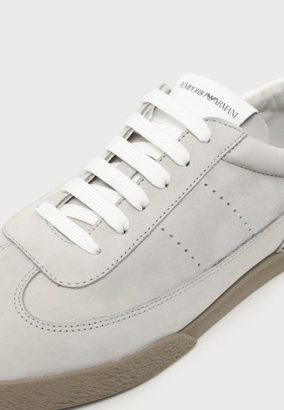 Gros plan sur une basket gris clair avec des lacets blancs, le logo Emporio Armani sur la languette et une semelle en caoutchouc beige texturée.