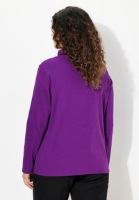 Lila Langarmoberteil aus einem weichen Stoff. Hat einen entspannten Schnitt mit geradem Saum und Ärmeln, kombiniert mit schwarzen Hosen.