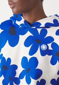 Sudadera con capucha con patrones de flores azules sobre un fondo blanco. Presenta una pequeña etiqueta circular azul con una cara sonriente.