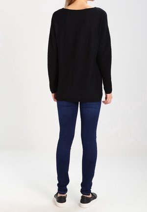 Maglietta nera a maniche lunghe con texture a coste sulle spalle, abbinata a jeans slim-fit blu scuro e scarpe nere slip-on.