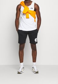 Bílý tílko s černým logem, černé šortky s bílou grafikou, oranžový svetr uvázaný kolem pasu a šedé tenisky se žlutými akcenty.