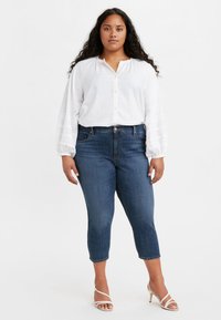 Chemise blanche boutonnée avec manches bouffantes, associée à un jean court bleu foncé et des sandales blanches à lanières. Tenue simple et décontractée.