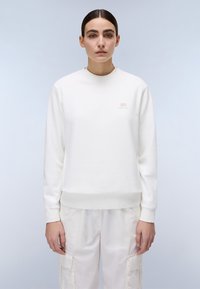 Vit sweatshirt med rund halsringning, ribbad mudd och nederkant, med en liten logo på bröstet. Matchad med ljusa cargobyxor.