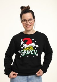 Černá mikina s kapucí, na které je kreslený Grinch v červené vánoční čepici, se zeleným nápisem "GRINCH" a bílými hvězdicovými akcenty.