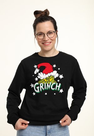 Svart sweatshirt med en tecknad Grinch i en röd julsnäppa, med grön text som säger "GRINCH" och vita stjärndetaljer.