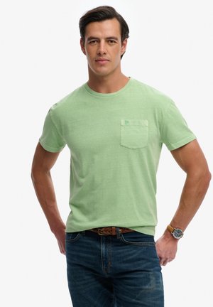 Mann trägt ein hellgrünes T-Shirt mit kurzer Ärmeln und Tasche, blaue Jeans, braunen geflochtenen Gürtel und Armbanduhr, steht mit den Händen in den Hüften.