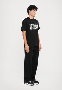 Svart bomulls-T-skjorte med stor hvit tekst "VERSACE COUTURE". Brukes med svarte denimbukser og svarte joggesko. Enkel, avslappet passform.