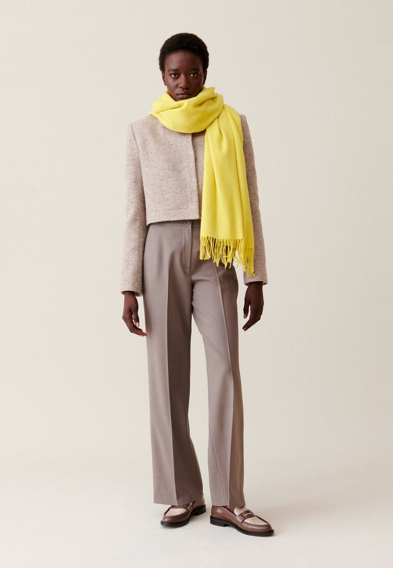 Stehende Person, die eine beige kurze Jacke, graue Hosen, braune Mokassins und einen leuchtend gelben Schal um den Hals trägt.