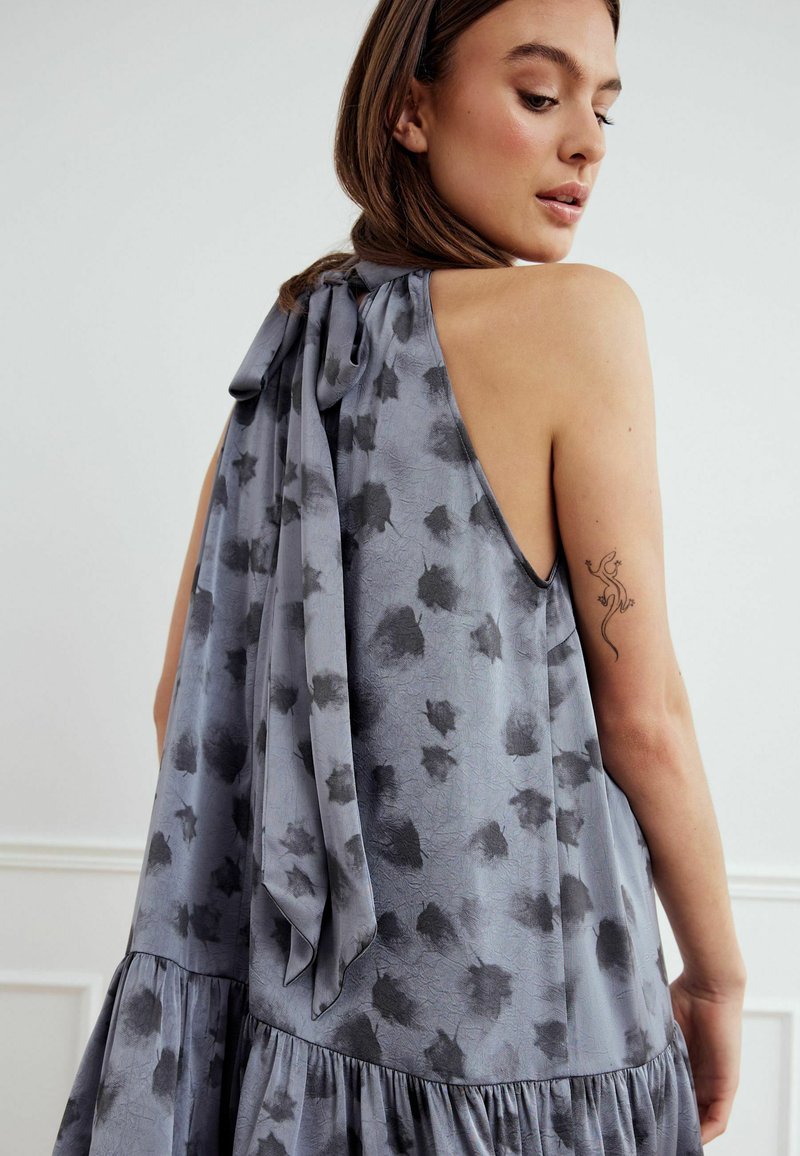 Siva haljina od tkanine sa slobodnim krojem, halter izrezom s mašnom i crnim apstraktnim uzorcima. Suknja ima višeslojni dizajn i teksturiranu površinu.