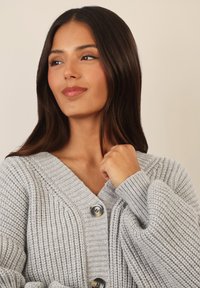 Cardigan lavorato a maglia grigio chiaro con scollatura a V, texture a costine, due grandi bottoni e maniche ampie.