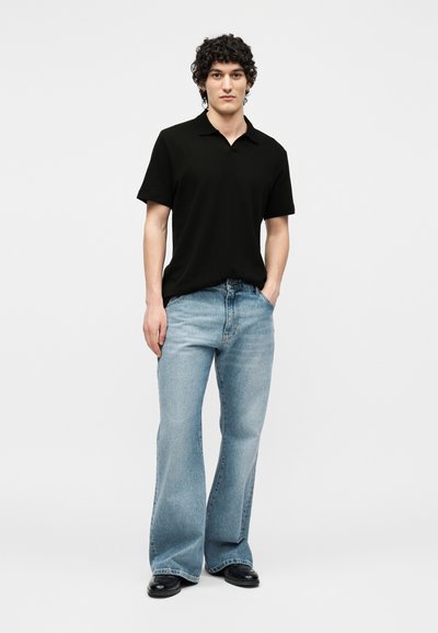 Jeune homme aux cheveux bouclés portant un polo noir à manches courtes, un jean large bleu clair et des chaussures noires, debout avec une main dans la poche.