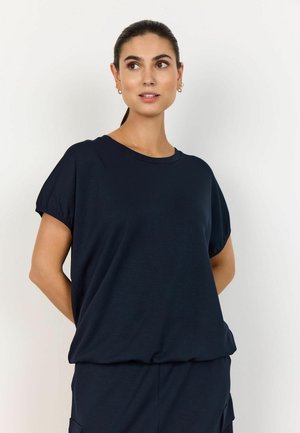 Kobieta z ciemnymi włosami związanymi do tyłu, ubrana w luźną, ciemnogranatową bluzkę z krótkim rękawem i pasujące spodnie, stojąca na jednolitym jasnym tle.