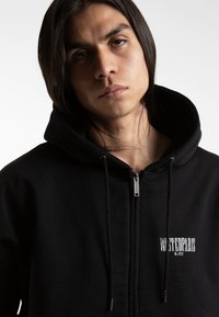 Čierna mikina s kapucňou na zips a šnúrkami, s vyšitým logom na hrudi. Mäkko hladký povrch s veľkou kapucňou a detailom na zips.