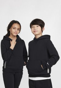 Due bambini indossano felpe nere abbinati con tasche a canguro. Le felpe hanno una finitura testurizzata e un piccolo logo Nike swoosh su una manica.