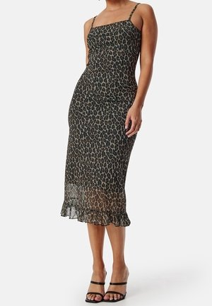 Kvinde iført en ærmeløs, leopardprint midi-kjole med tynde stropper og flæsekant, kombineret med sorte, åbne højhælede sandaler.