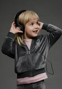 Veste zippée en velours gris avec des poignets en ribbed noirs, associée à une chemise rose clair en dessous. L'enfant porte des écouteurs noirs et sourit.