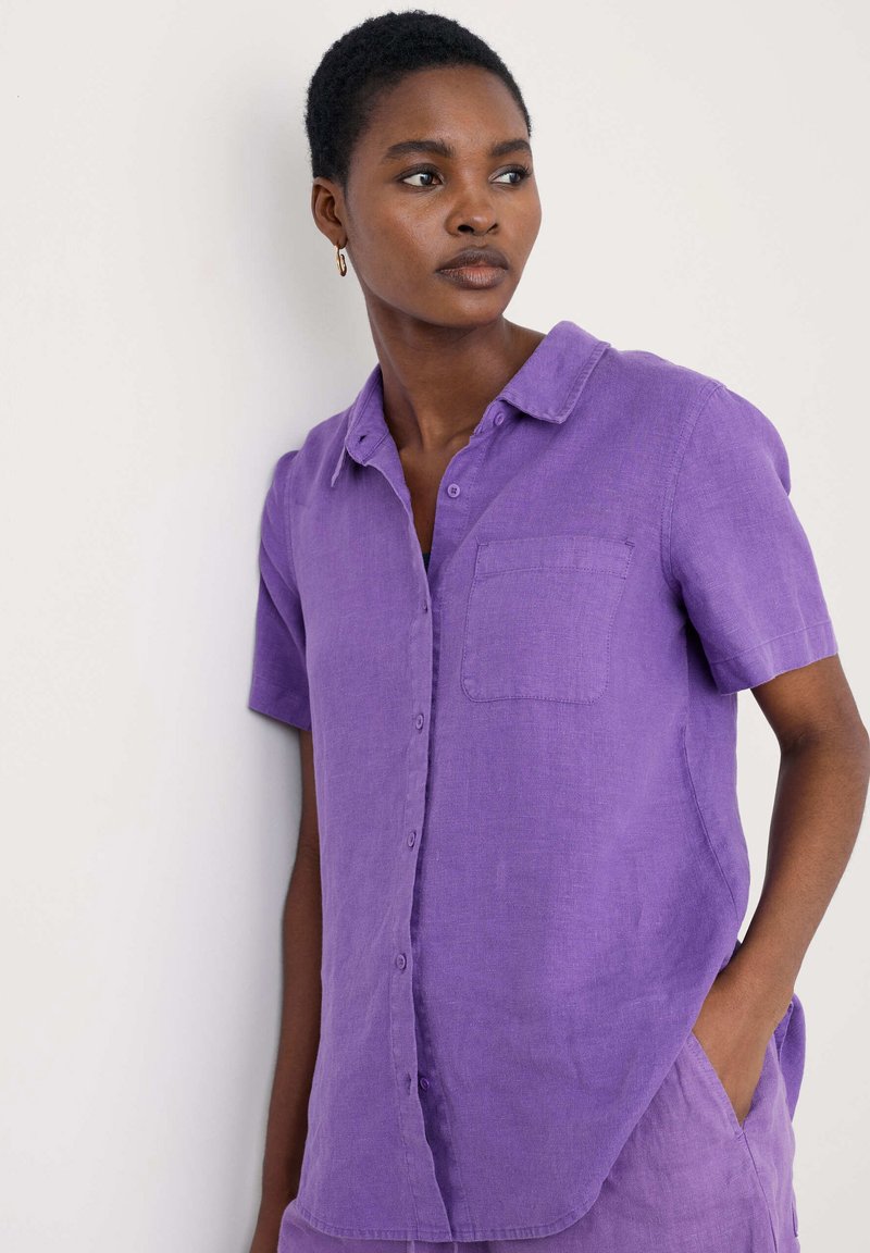 Violet trumpetas rankovėmis marškiniai, pagaminti iš lino, su mygtukų užsegimu priekyje, krūtinės kišenė ir laisvas siluetas. Sklandus tekst�ūra ir vienspalvė.