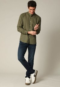 Olijfgroene overhemd met een mandarinokraag, gecombineerd met donkerblauwe jeans en witte sneakers met zwarte accenten.