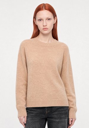 Jeune femme aux cheveux roux lisses portant un pull beige à manches longues et un jean foncé, debout devant un fond clair uni.