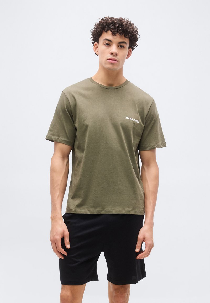 Νεαρός άνδρας με κατσαρά μαλλιά που φοράει πράσινο ελιάς μπλουζάκι Jack & Jones και μαύρο σορτς, στέκεται με ουδέτερη έκφραση.