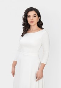 Weißes Kleid mit langen Ärmeln, enganliegendem Oberteil und fließendem Rock. Verfügt über einen runden Ausschnitt und eine glatte Textur. Elegantes und minimalistisches Design.