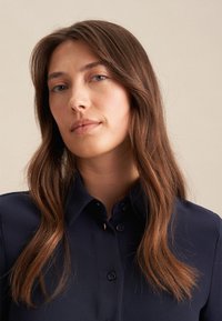 Navyblaues Hemd mit Knöpfen, das einen klassischen Kragen, eine glatte Textur und subtile Steppdetails aufweist, kombiniert mit langen Haaren, die das Gesicht umrahmen.