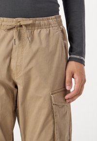 Beige cargo pants featuring a drawstring waist, side pocket, and relaxed fit. Fabric appears lightweight with a smooth texture.