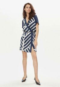 Abito avvolgente a fantasia blu navy e crema con maniche corte, caratterizzato da un profondo scollo a V, spacco laterale e cintura in tessuto. Tacchi neri eleganti completano il look.