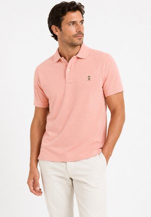 Homme portant un polo rose clair à manches courtes avec un petit emblème sur la poitrine et un pantalon blanc, debout avec une main dans la poche.
