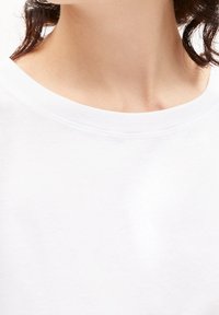 Närbild på en persons nacke och övre bröst som bär en enkel vit T-shirt med rund halsringning, med mörkt hår synligt.