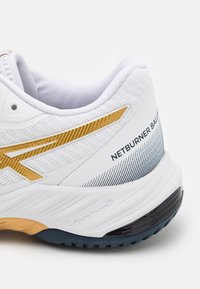 Baskets blanches de sport avec un dessus en maille texturée, une bande d'accent dorée, le texte "NETBURNER BALL" et une semelle extérieure contrastante noire et bleue.