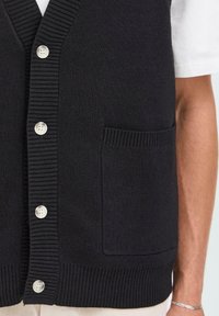 Zwarte gebreide knoopvest met een patch pocket, gedragen over een witte t-shirt met korte mouwen, waarbij een deel van een tan arm en een zilveren armband zichtbaar is.