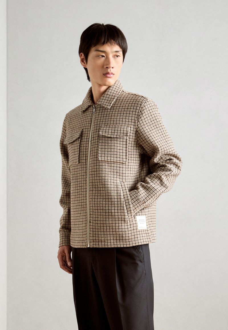 Chaqueta marrón de cuadros con cremallera en el frente, dos bolsillos en el pecho y un acabado texturizado, combinada con pantalones negros.