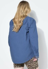 Modrá košile s dlouhým rukávem a volným střihem, s manžetami na knoflíky a detailním akcentem šití. Na sobě má šortky s leopardím potiskem.