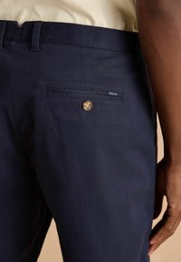 Pantaloni din bumbac marină, având un buzunar posterior cu buton și un etichetă de marcă vizibilă. Țesătura pare netedă, cu un ușor luciu.