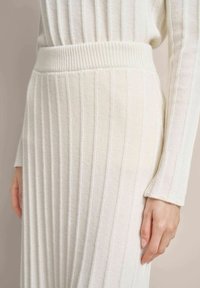 Knit cream skirt with vertical ribbed texture and elastic waistband, featuring long sleeves from a matching top, hand resting on the fabric.