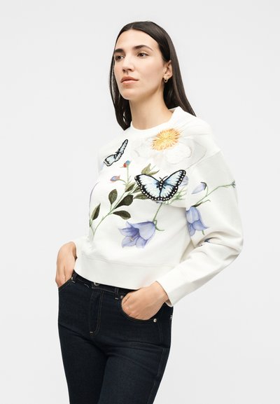 Γυναίκα με σκούρα μαλλιά που φοράει λευκό φούτερ με κεντητές πεταλούδες, λουλούδια και πράσινα φύλλα, συνδυασμένο με σκούρο τζιν.