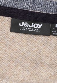 Brede gebreide stof in lichtbeige met subtiele textuur, voorzien van een donkergrijze rib trim en twee zwarte labels met witte tekst.