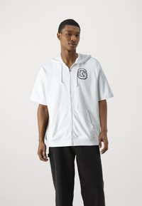 Bijela dukserica s kapuljačom i kratkim rukavima, sa crnim izvezenim "G" logotipom na prsima i prednjim džepovima.