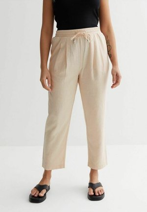 Kobieta w beżowych lnianych spodniach z wysokim stanem, z elastycznym sznurkiem i czarnych sandałach na platformie, stojąca na jednolitym jasnym tle.