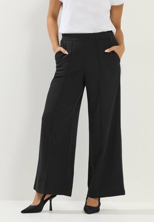 Frau trägt schwarze, weit geschnittene Hosen mit vorderen Nähten, kombiniert mit schwarzen Slingback-Pumps mit hohem Absatz und einem weißen, in die Hose gesteckten Oberteil. Hände in den Taschen.