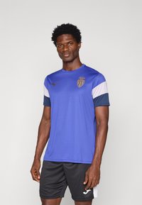 Modrá športová košeľa s bielymi a tmavomodrými manžetami, s logom na hrudi a okrúhlým golierom. Čierne šortky dopĺňajú outfit.