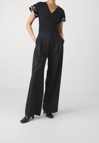 Mustapainen V-pääntie toppi, jossa on rypytetty yksityiskohta ja lyhyet hihat, yhdistettynä korkeavyötäröisiin, leveälahkeisiin mustiin housuihin ja mustiin korkokenkiin.