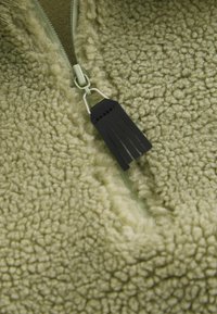 Roheline fliisjope lähivõte, millel on tekstuuriga pind, kontrastne tõmblukukäpp ja tõmbluku narmasdetail.