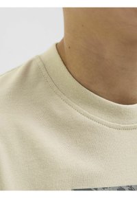 Beżowa bawełniana koszulka z okrągłym dekoltem, z teksturowanym splotem i subtelnym detalem przeszycia. Górna część klatki piersiowej zdobi abstrakcyjna grafika.