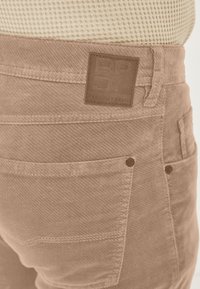 Beige Cord-Hosen mit strukturierter Oberfläche, mit einem quadratischen Lederpatch, Knopfakzenten und mehreren Taschen in Taillennähe.