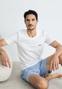 T-shirt en coton blanc avec le logo "BOSS" en noir, associé à un short bleu clair. Texture lisse, col rond, coupe décontractée. Fond clair.