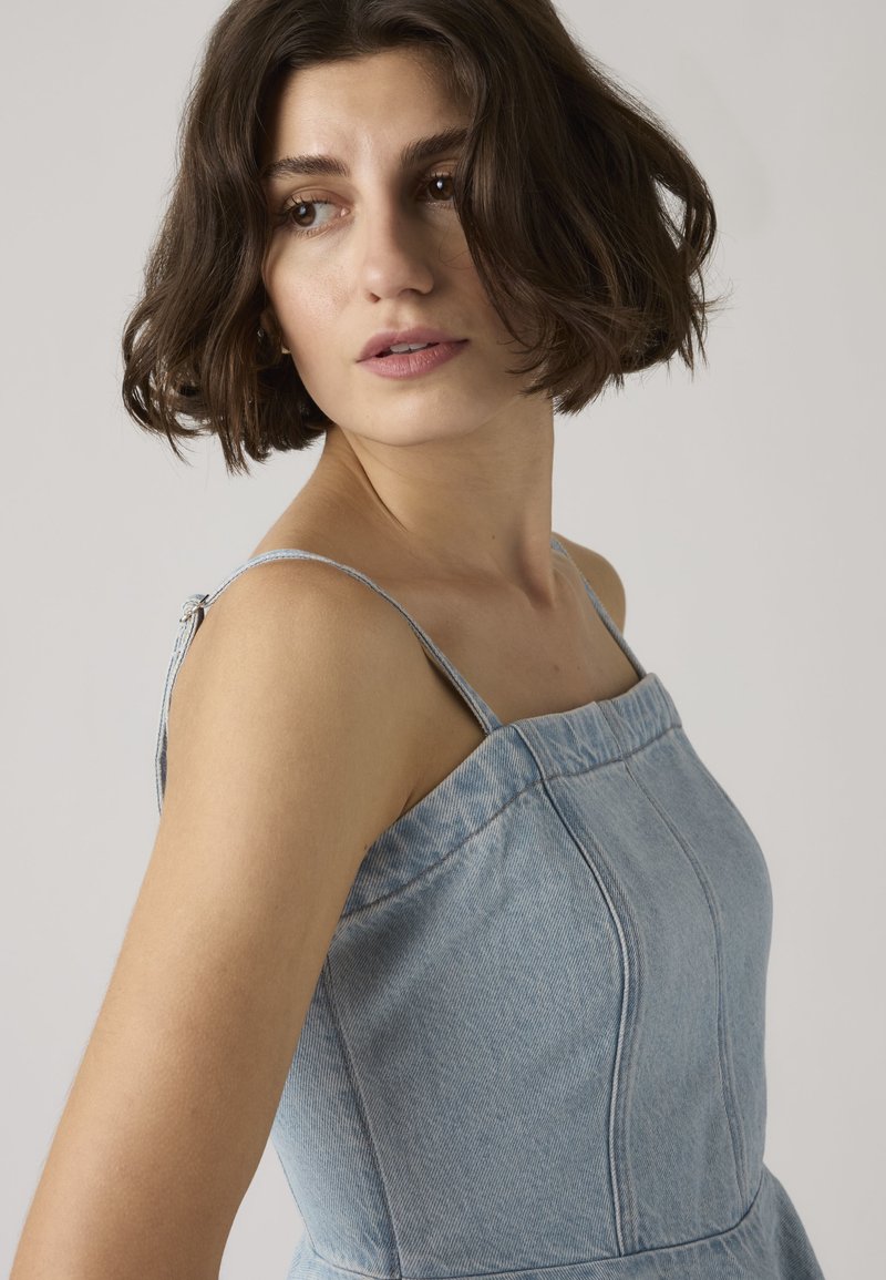 Jonge vrouw met kort bruin haar, gekleed in een mouwloze lichtblauwe spijkerjurk, die iets naar links kijkt tegen een effen achtergrond.