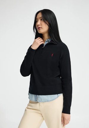 Kobieta z długimi czarnymi włosami, ubrana w czarny sweter na zamek, na koszulę dżinsową i beżowe spodnie, stojąca na tle jasnego, jednolitego tła.