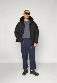 Crna puffer jakna, siva majica, mornarsko plave cargo hlače i crne tenisice. Uključuje sunčane naočale i dva džepa na hlačama.